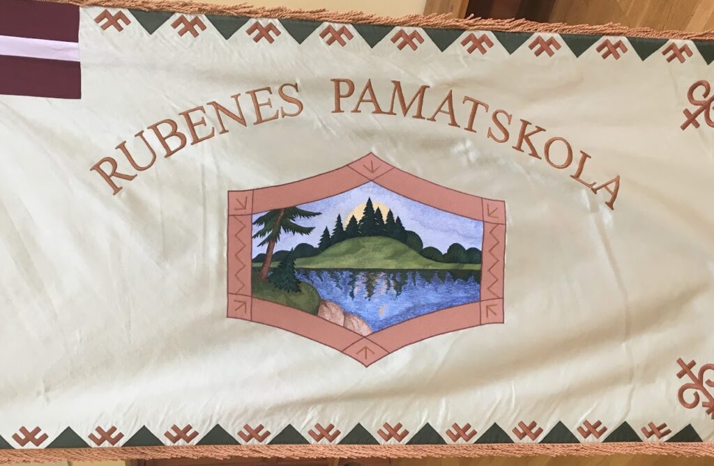 Gaišas krāsas auduma karogs ar izšuvumu, kura augšpusē puslokā izšūts teksts "RUBENES PAMATSKOLA". Karoga centrā ir rombveida rāmis ar dabas ainavu: ezeru priekšplānā un mežainu pakalnu fonā ar uzlecošu sauli. Karoga apmales rotātas ar rakstu no  zaļiem trīsstūriem un rūsas krāsas nacionālajiem ornamentiem. Karogam ir bārkstis gar malām. Augšējā kreisajā stūrī redzams neliels Latvijas valsts karogs.