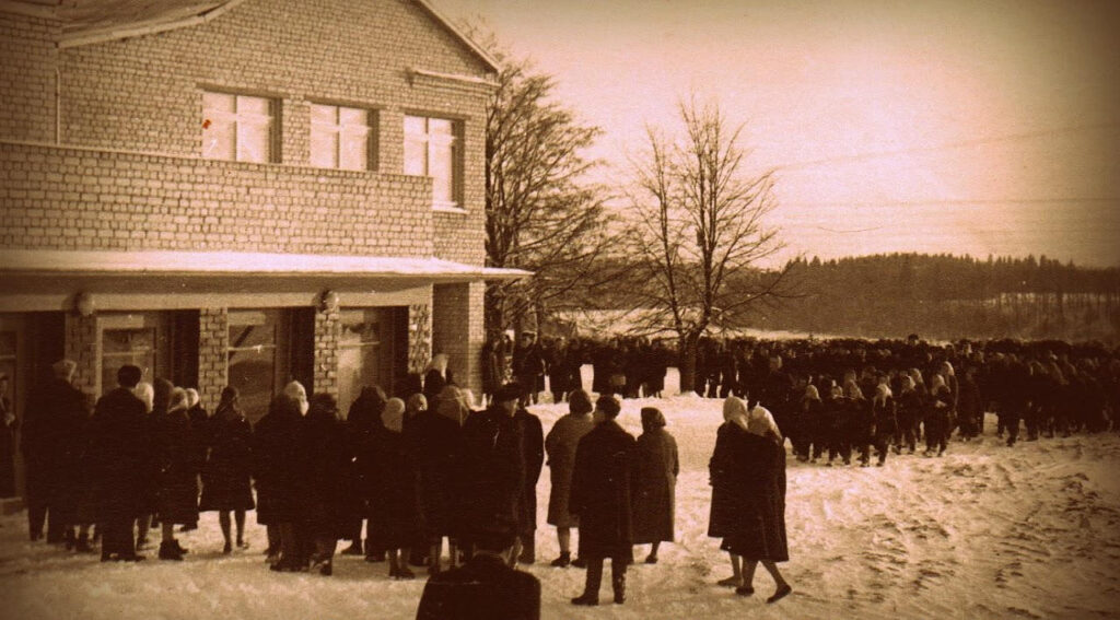 Sena, ruda toņa fotogrāfija, kurā ziemā, sniegā pie jaunuzceltās divstāvu ķieģeļu skolas ēkas svinīgā pasākumā pulcējas liels cilvēku pūlis.