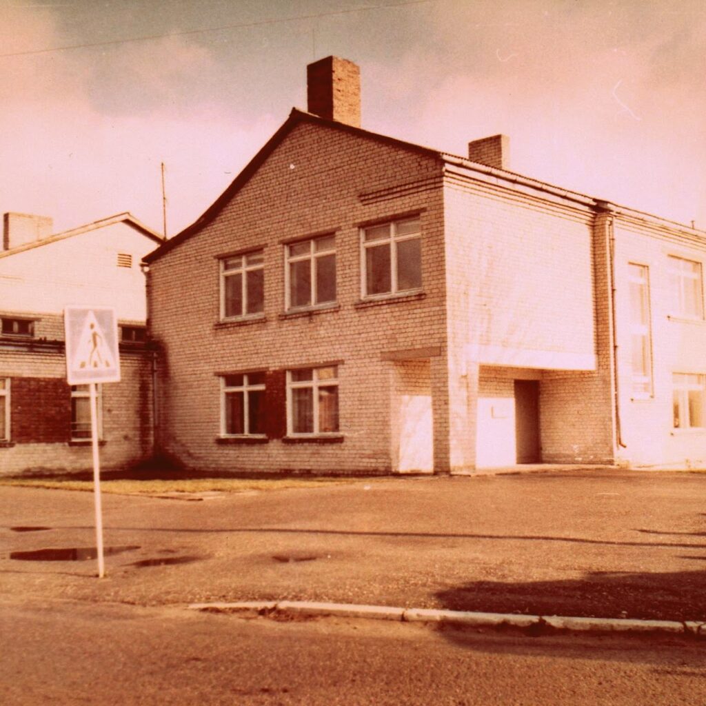 Sena, krāsaina fotogrāfija ar divstāvu ēku no baltiem ķieģeļiem - Rubenes skola. Priekšplānā pie asfaltēta ceļa malas uzstādīta ceļa zīme "Gājēju pāreja".