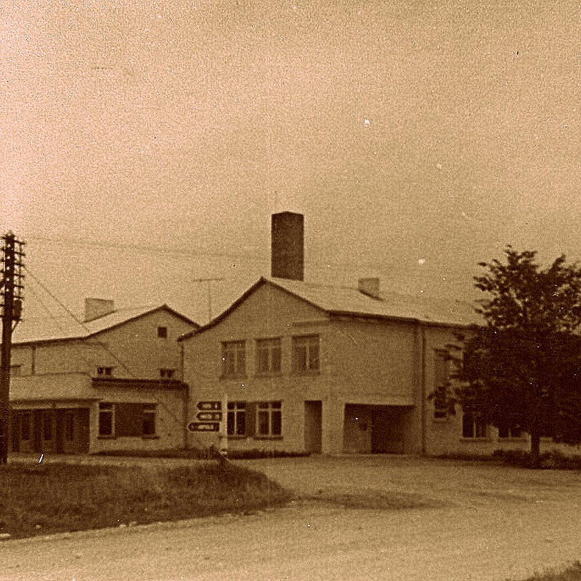 Vēsturiska fotogrāfija. Tālajā plānā redzama baltu ķieģeļu ēka - Rubenes skola. Priekšplānā ceļš ar grunts segumu, virzienu rādītāji un telefona līnijas stabs.