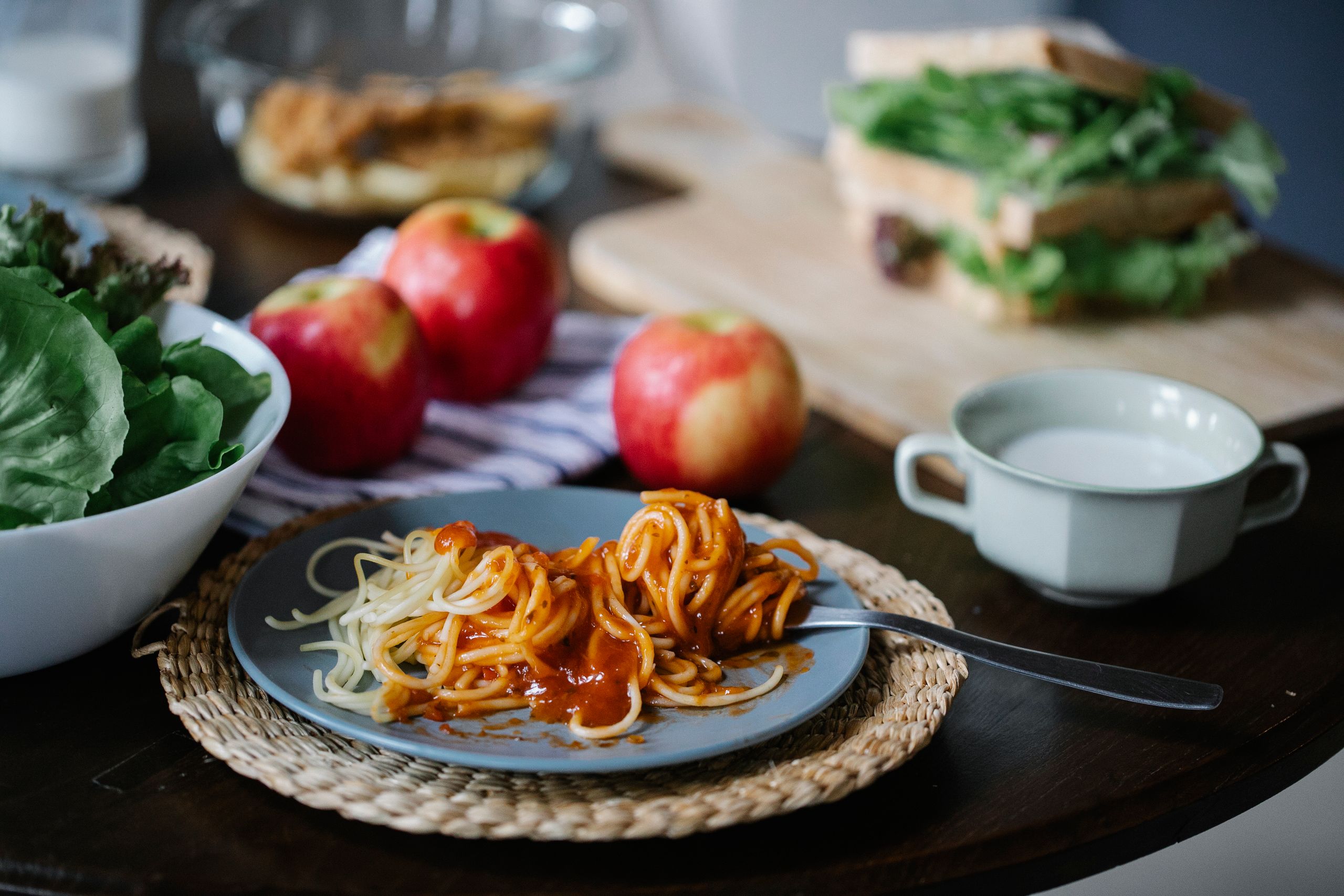 Appetizing spaghetti with tomato sauce on plate with fork placed on table with apples and greens in kitchen with blurred background